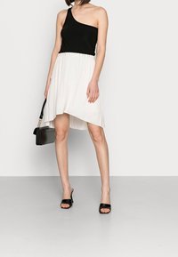 Schwarzes einseitiges Oberteil kombiniert mit einem weißen asymmetrisch gefalteten Rock. Das Model hält eine schwarze Handtasche und trägt schwarze offene Schuhe.