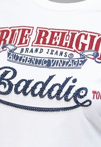 Biała bawełniana koszulka z czerwoną i niebieską teksturą z kryształkami, na której widnieje napis "True Religion Brand Jeans Authentic Vintage Baddie."