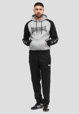 Homme debout les mains dans les poches, portant un sweat à capuche Lonsdale London gris et noir, un pantalon de survêtement Lonsdale London noir, et des baskets noires.