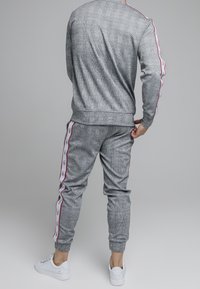 Grau gemusterter Jogginganzug mit roten Seitenstreifen, aus strukturiertem Stoff. Verfügt über elastische Bündchen und eine figurbetonte Passform, kombiniert mit weißen Sneakers.