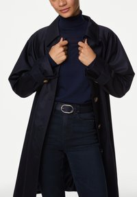 Trinchera impermeable en color navy con cuello, que presenta botones grandes. Llevado sobre un cuello de tortuga navy y jeans de cintura alta con una hebilla ovalada plateada.
