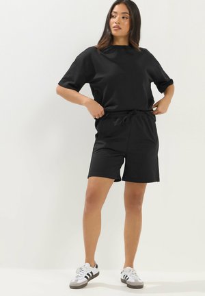 Vrouw die een zwart T-shirt met korte mouwen en zwarte shorts aanpast, draagt witte sneakers met zwarte strepen, staand tegen een witte achtergrond.
