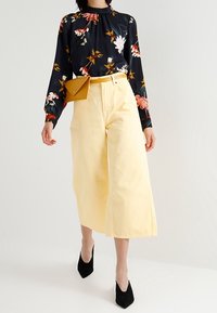 Blusa floreale a maniche lunghe blu navy con accenti rossi, rosa e verdi; pantaloni gialli a vita alta e a gamba larga; abbinati a tacchi neri e a una borsa senape.