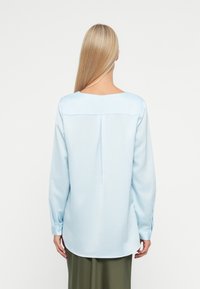 Svetlo modra bluzica z dolgimi rokavi, zaobljenim izrezom in naborkom na hrbtni strani. Mehka tekstura in rob, ki se rahlo ukrivi ob straneh.