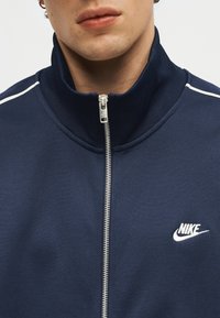 Κοντινό πλάνο ενός ατόμου που φοράει μπλε-ναυτικό τζάκετ Nike με φερμουάρ, με λευκό λογότυπο και λευκές ρίγες στους ώμους.