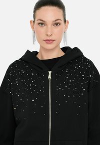 Une femme aux cheveux tirés en arrière porte un sweat à capuche noir orné de paillettes brillantes, arborant une expression neutre.