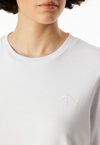 Vit bomullströja med ribbat rundhals och subtil broderad logotypdetalj på bröstet. Slät textur, avslappnad passform.