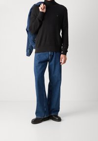 Person trägt schwarzen strukturierte Rollkragenpullover, lockere blaue Jeans und schwarze Schuhe, hält eine blaue Jeansjacke über der Schulter.