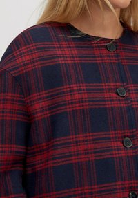 Rood en marineblauw geruite button-up jas van een zachte, textuurstof met een ronde halslijn en vier zwarte knopen.