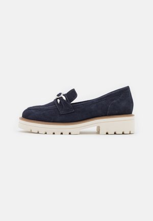 Navy suede loafers feature a low, chunky white sole and a decorative bow accent. The design is sleek with contrasting stitching details.