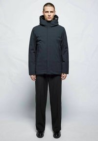 Chaqueta aislante negra con cuello alto y capucha ajustable, combinada con pantalones negros holgados y zapatos negros brillantes.