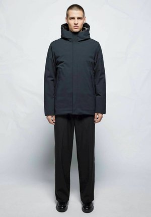Zwarte geïsoleerde jas met hoge kraag en verstelbare capuchon, gedragen met losse zwarte broek en glanzende zwarte schoenen.