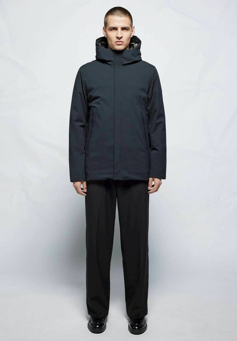 Chaqueta aislante negra con cuello alto y capucha ajustable, combinada con pantalones negros holgados y zapatos negros brillantes.