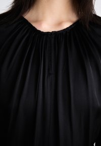 Personne portant un chemisier noir à encolure froncée, vue des épaules à la clavicule, avec des cheveux lisses et foncés encadrant le haut de l'image.