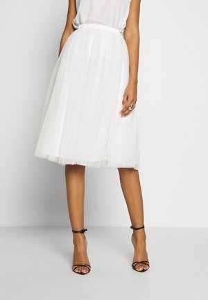 Vrouw draagt een witte geplooide midi-rok met een gerimpelde tailleband en zwarte sandalen met hoge hakken en bandjes, staand tegen een effen achtergrond.
