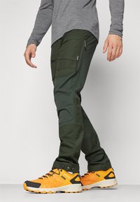 Ciemnozielone spodnie cargo z wieloma kieszeniami, detalami na suwak oraz zwężanym krojem, w połączeniu z jasn Yellowimi butami trekkingowymi.