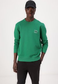 Felpa verde con polsini e orlo a coste, caratterizzata dal logo bianco "Tommy Hilfiger" sul petto. Indossata con pantaloni neri.