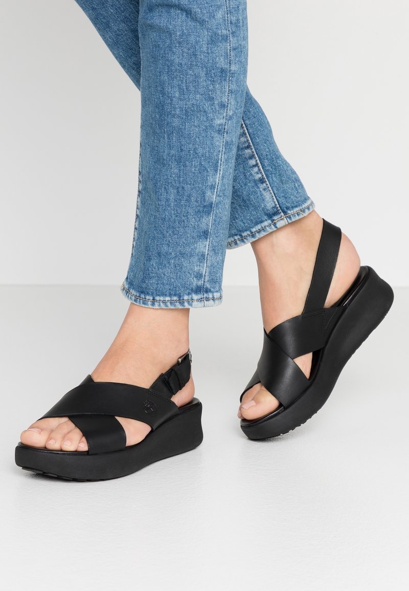 Pieds portant des sandales compensées noires à brides croisées et un jean en denim bleu sur un fond clair uni.