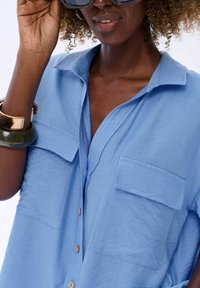 Light blue button-up shirt made of textured fabric, featuring two front pockets and a classic collar. Gold and green bangle accessories visible.