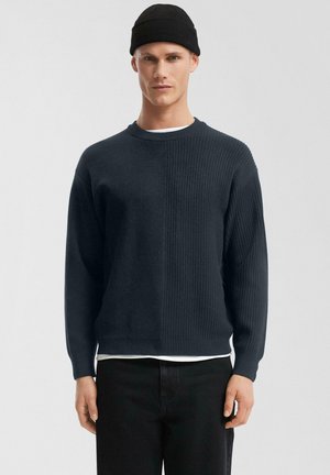 Mann trägt schwarze Strickmütze, dunkelblauen gerippten Pullover über weißem Hemd und schwarze Hose, steht vor einem schlichten hellen Hintergrund.