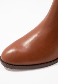 Bota de tobillo de cuero marrón con textura lisa, punta puntiaguda y suela de goma oscura con detalles texturizados a lo largo del borde.