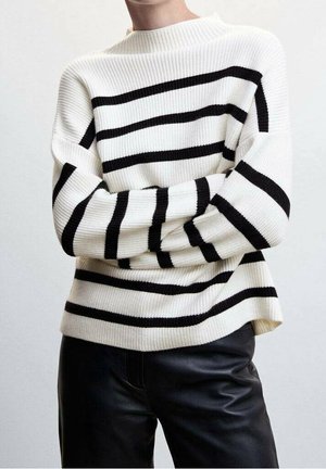 Personne portant un pull en maille blanc à rayures horizontales noires et un pantalon en cuir noir, bras croisés sur la poitrine.