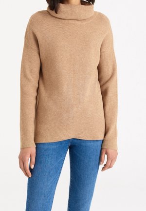 Suéter de punto acanalado beige con cuello de barco, manga larga y corte relajado. Detalles de costura visibles y textura suave. Combinado con vaqueros azules.