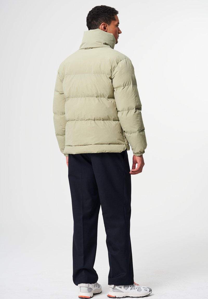 Hellgrüne Steppjacke mit hohem Kragen, gesteppter Optik und elastischen Bündchen, getragen über navyfarbener Hose und weißen Sneakern.