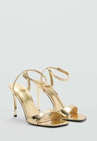 Zlaté sandále na vysokom opätku s otvorenou špičkou, s tenkým remienkom okolo členku, lesklým kovovým povrchom a elegantným stiletto opätkom.