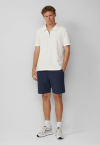 Herren-Polohemd in Creme mit Reißverschluss-Kragen, kombiniert mit marineblauen Shorts. Das Modell trägt graue Sneakers und weiße Socken. Einfaches, modernes Design und leichter Stoff.