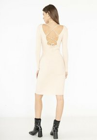 Robe beige côtelée avec des manches longues et un dos plongeant doté de bretelles croisées et d'un nœud, accompagnée de bottines noires brillantes à la cheville.