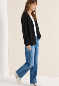 Černý žebrovaný kardigan přes bílou košili, v kombinaci s světlomodrými džínami s rovnými nohavicemi a bílými teniskami s červenými detaily.