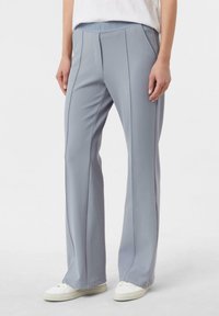 Osoba ubrana w jasnoniebieskie spodnie o prostych nogawkach z przednimi przeszyciami, kieszeniami po bokach i białe trampki, stojąca na jednolitym tle.