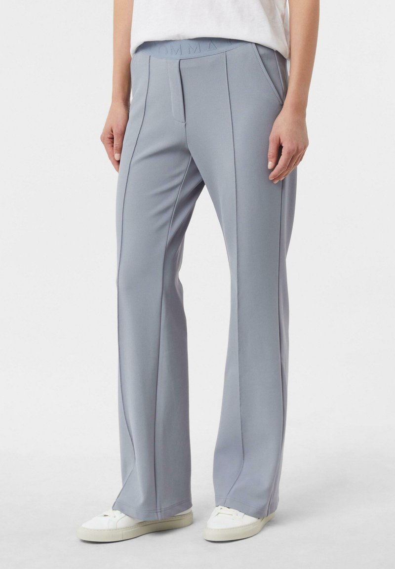 Osoba ubrana w jasnoniebieskie spodnie o prostych nogawkach z przednimi przeszyciami, kieszeniami po bokach i białe trampki, stojąca na jednolitym tle.