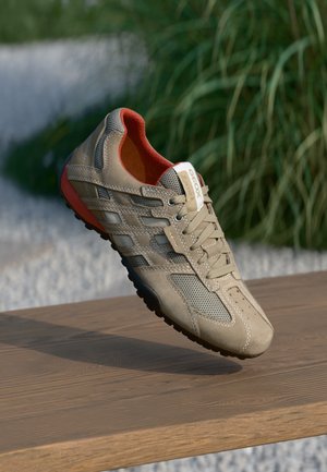 Baskets décontractées beige et grises avec intérieur rouge et semelle noire reposant sur une surface en bois, avec un arrière-plan flou de verdure.