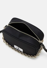 Bolso cruzado negro con dos cremalleras, acento de cadena plateada y una textura suave. Presenta un parche de logo visible en un lado.