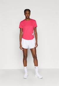 Ροζ αθλητικό μπλουζάκι με κοντά μανίκια, στρογγυλεμένη ραφή και λευκό λογότυπο. Συνδυάζεται με λευκό σορτς και λευκά αθλητικά παπούτσια, φορεμένα με λευκές κάλτσες.