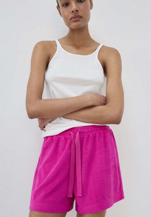 Mladá osoba stojaca so založenými rukami, oblečená v biele tielko a jasne ružové šortky so šnúrkou na jednofarebnom pozadí.