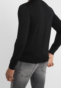 Czarny sweter z długim rękawem i okrągłym dekoltem, z prążkowanymi mankietami i dołem, pokazany z tyłu na osobie w szarych dżinsach.