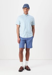 Chemise à manches courtes bleu clair à boutons, shorts bleus, chaussettes blanches, mocassins marron et casquette bleue. Tenue d'été simple et décontractée.