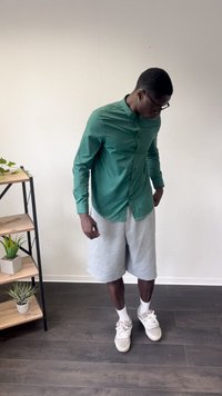 Grünes Langarmhemd mit Knopfleiste, kombiniert mit grauen Shorts. Getragen mit weißen Socken und hellen Sneakers. Einfarbiger Hintergrund.