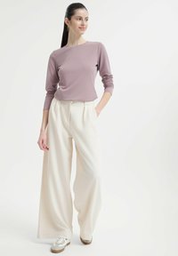 Helllila langärmliges Oberteil, kombiniert mit cremefarbenen weit geschnittenen Hosen. Das Outfit zeichnet sich durch eine glatte Textur und einen lässigen Schnitt aus, mit einem minimalistischen Design.