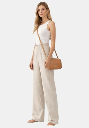 Donna che indossa un top bianco senza maniche, pantaloni beige a gamba larga, sandali bianchi e una borsa a tracolla color cuoio, in piedi contro uno sfondo semplice.