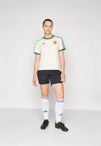 Vit och grön kortärmad tröja med röd logotyp, kombinerad med svarta shorts, vita strumpor med blå ränder och svarta fotbollsskor.