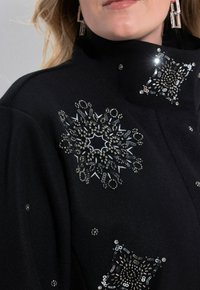 Gros plan sur une personne portant un manteau noir orné de motifs complexes de flocons de neige en perles et sequins argentés sur le col et l'épaule.