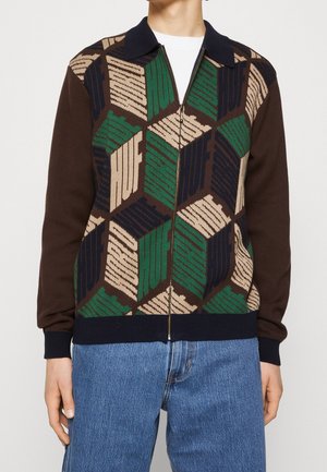 Osoba nosząca sweter na zamek z geometrycznymi wzorami w zielonym, beżowym i granatowym kolorze oraz powtarzającym się napisem "HUF", w połączeniu z niebieskimi dżinsami.