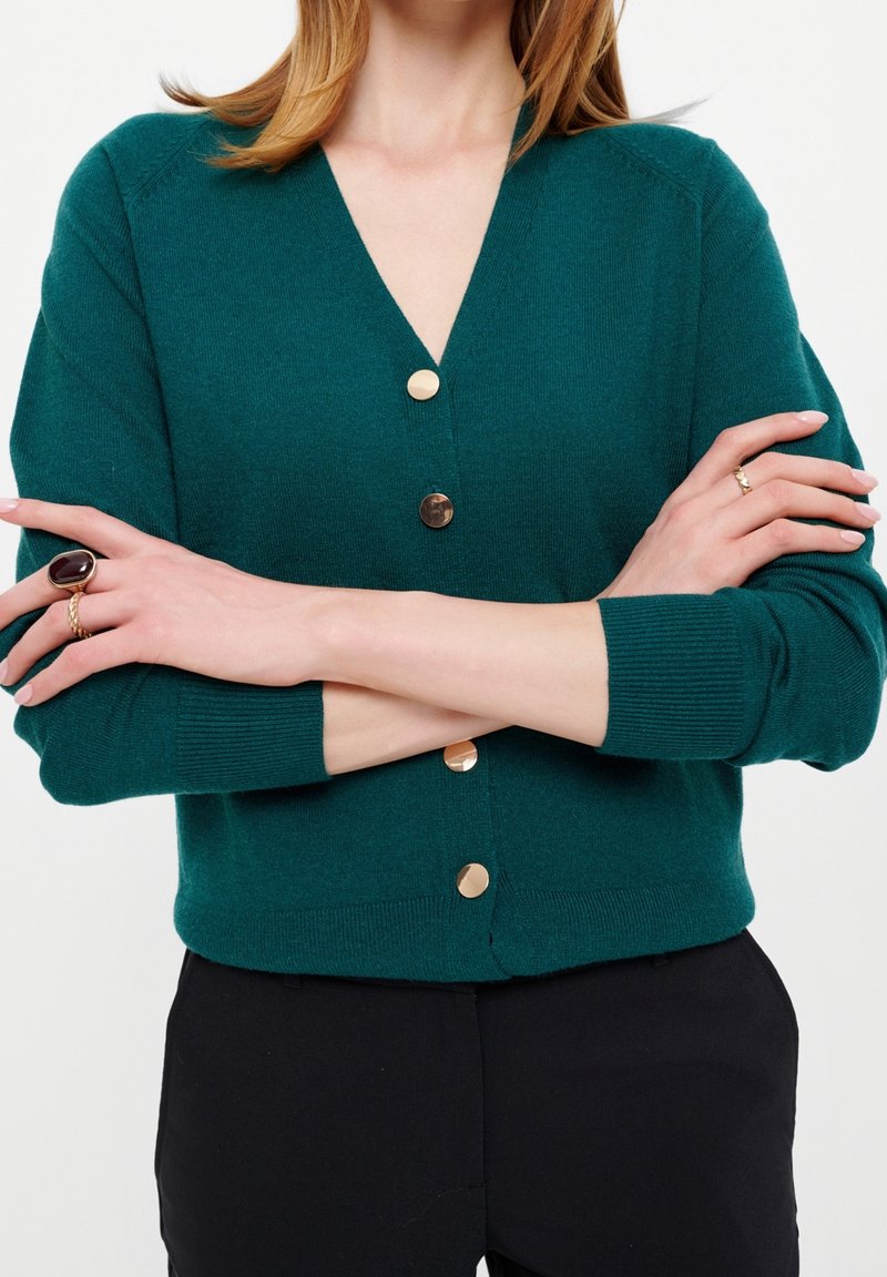 Femme portant un cardigan vert foncé avec des boutons dorés et un pantalon noir, bras croisés, affichant des bagues aux deux mains.