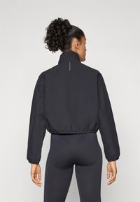 Cropped black windbreaker jacket with a high collar, elasticated cuffs and hem, featuring a reflective strip at the back.