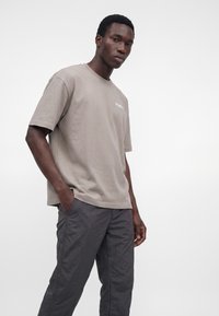 Jeune homme portant un t-shirt DropSeat beige et un pantalon gris foncé, debout avec une main dans la poche devant un fond blanc uni.