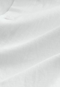 Nahaufnahme eines weißen, strukturierten Stoffs mit einem einzelnen durchscheinenden Knopf, der in der Nähe der oberen linken Ecke angenäht ist.